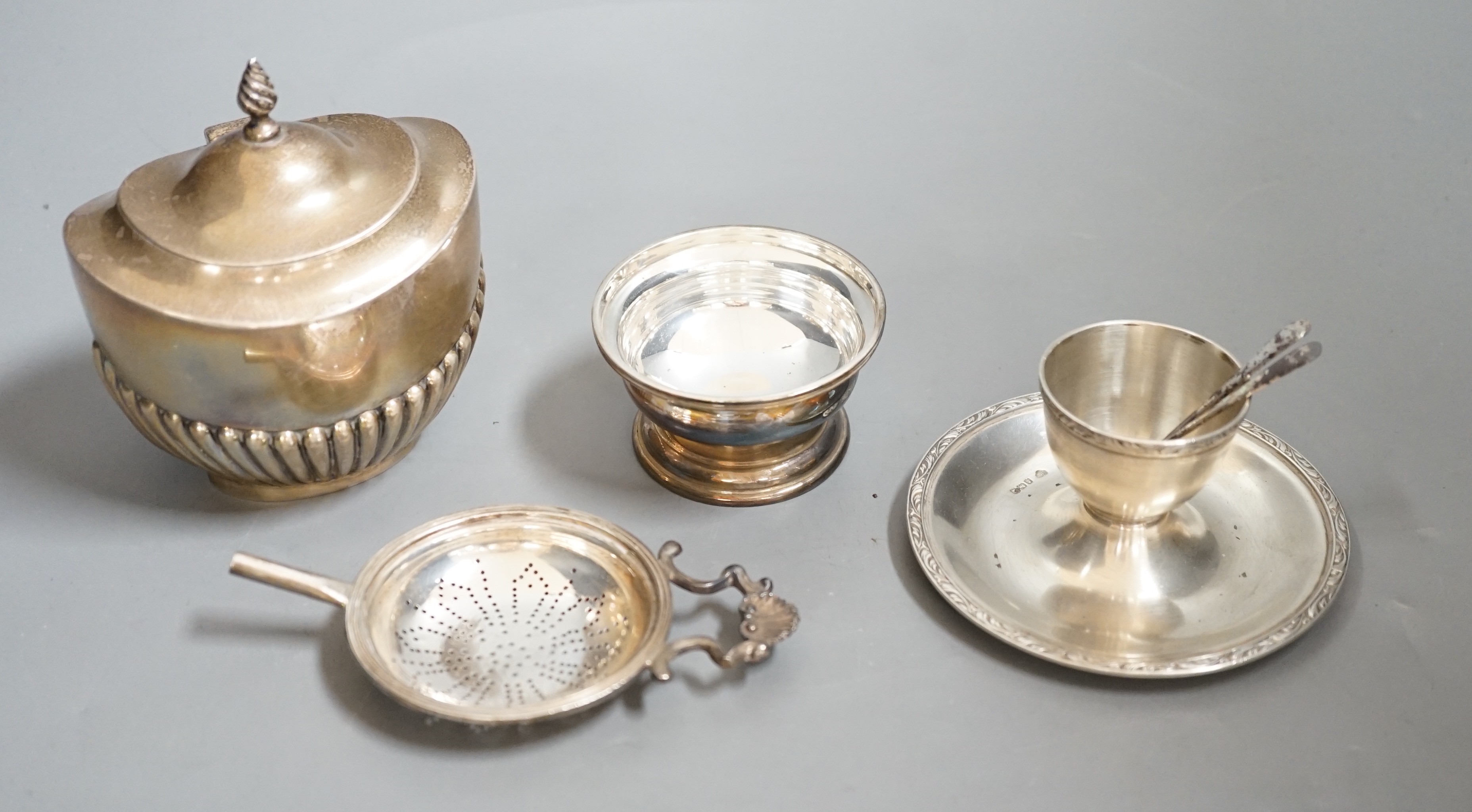 A late Victorian silver half-fluted tea caddy, Birmingham, 1900, a silver strainer on stand, a silver egg cup stand and a pair of silver server spoons.